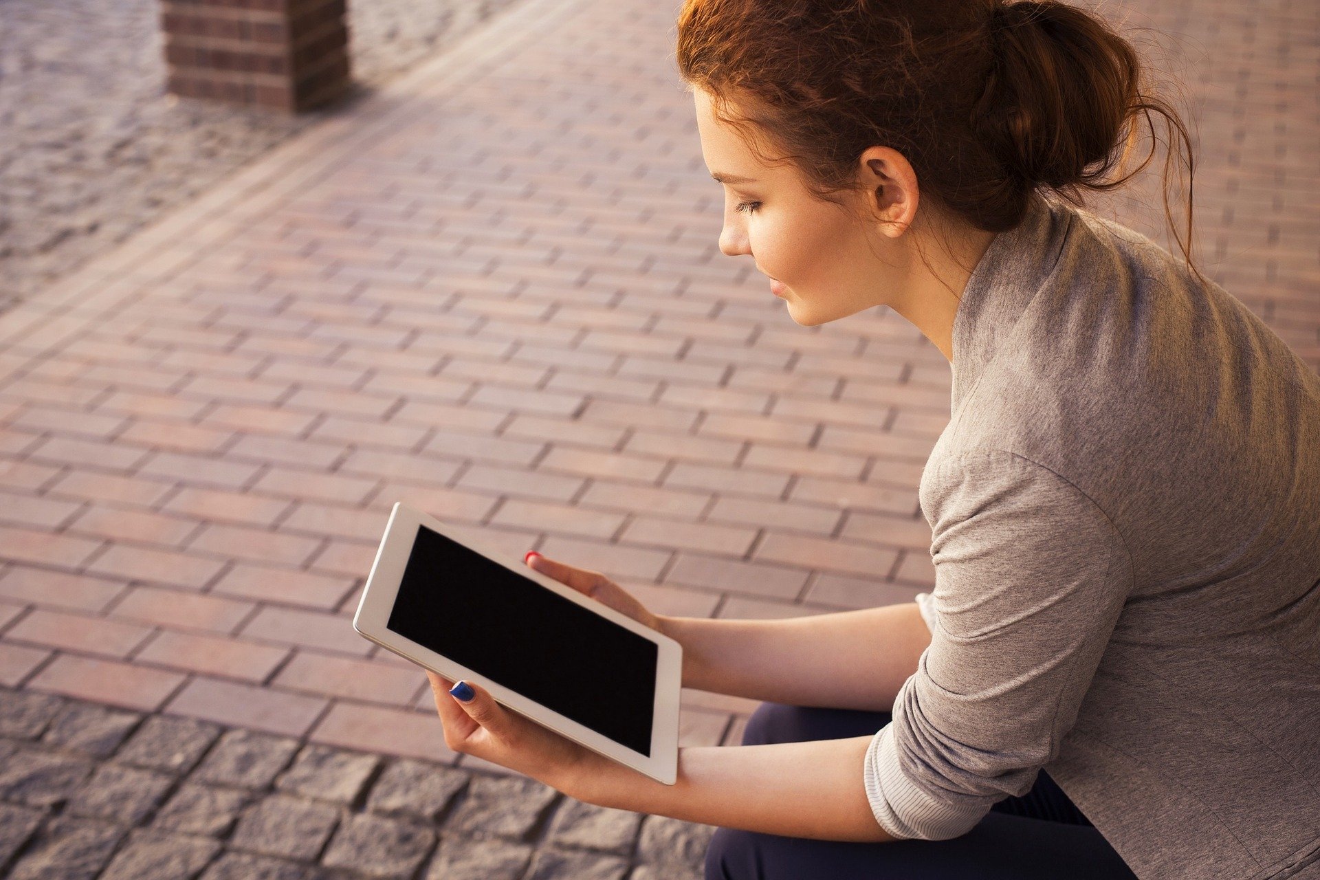  A young woman is reading a novel on her tablet while sitting on a bench.
