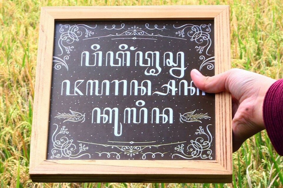  A wooden frame with a Javanese quote about life written in white letters on a black background, held up by a hand in a field of rice.